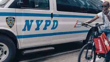 Food delivery cyclist breaking NYC bike laws next to police van.