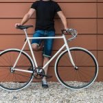 Image of a man wearing jean shorts holding a white fixed gear bike.