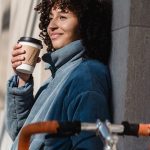Image of a woman with a blue jacket sitting and drinking coffee next to her bicycle. Source: Unsplash