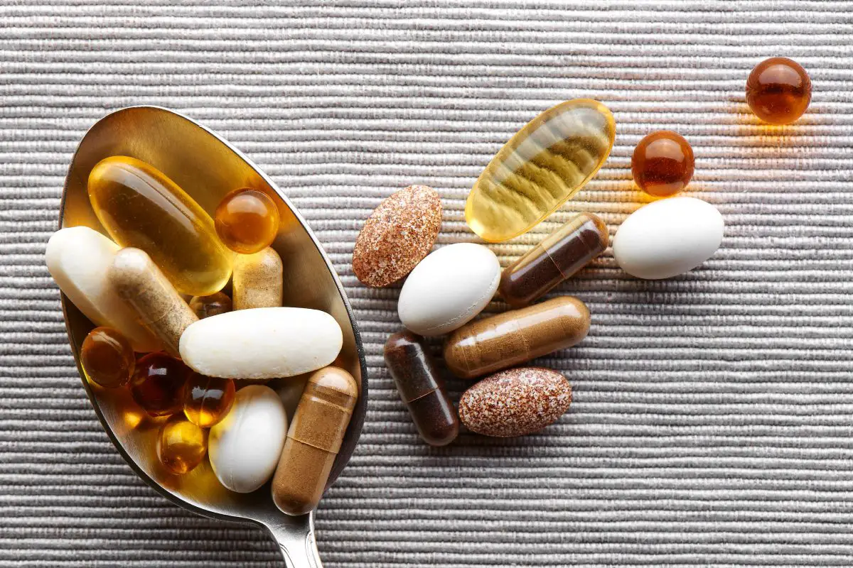 Close up tablespoon filled with various dietary supplements tablets and vitamins on a gray fabric background. Source: adobe stock