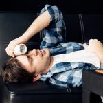 Image of a man lying on sofa and holding bottle near head after party. Source: Adobe Stock