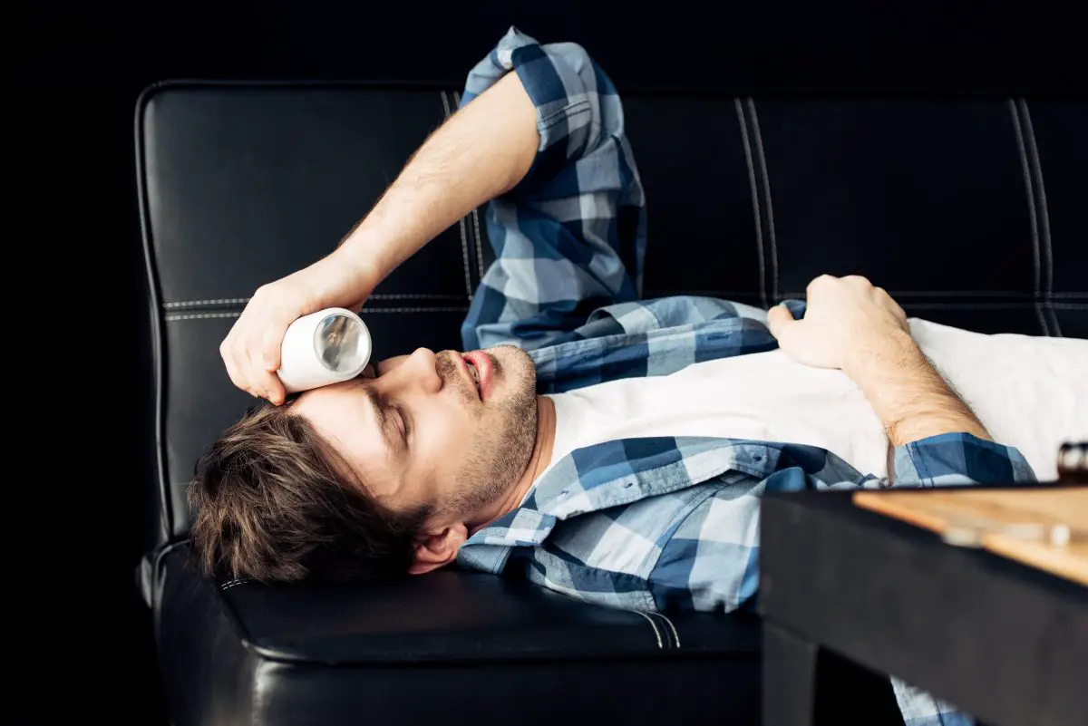 Image of a man lying on sofa and holding bottle near head after party. Source: adobe stock