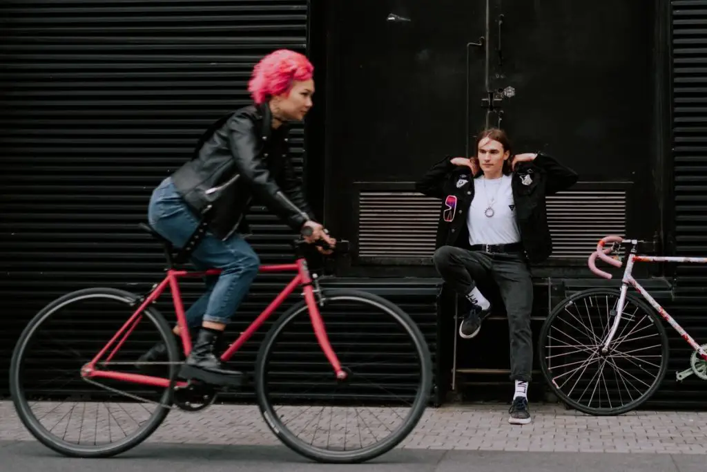 Image of a woman riding a fixie bike and a man sitting beside a bike. Source: pexels